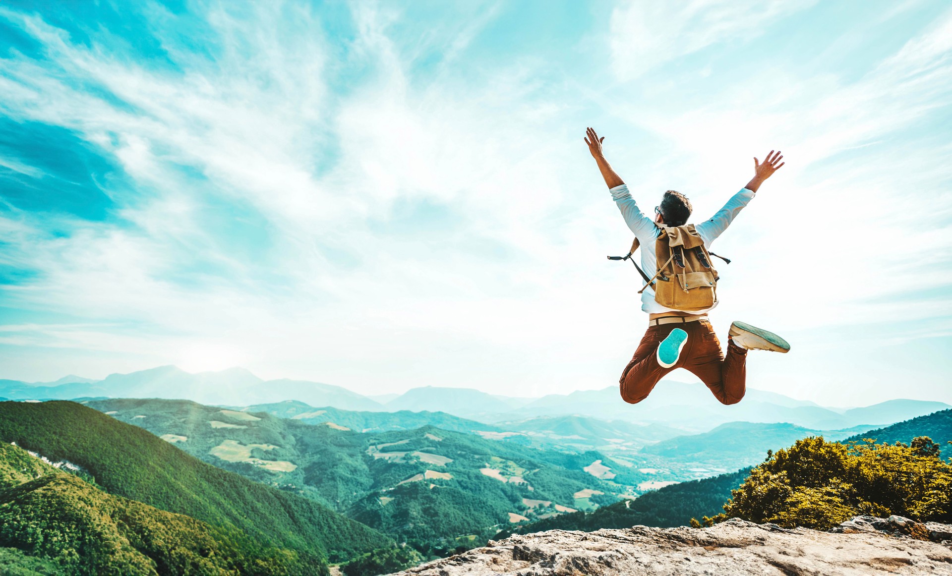 Happy man with backpack jumping on top of the mountain - Delightful hiker with arms up standing over the cliff - Sport and travel life style concept